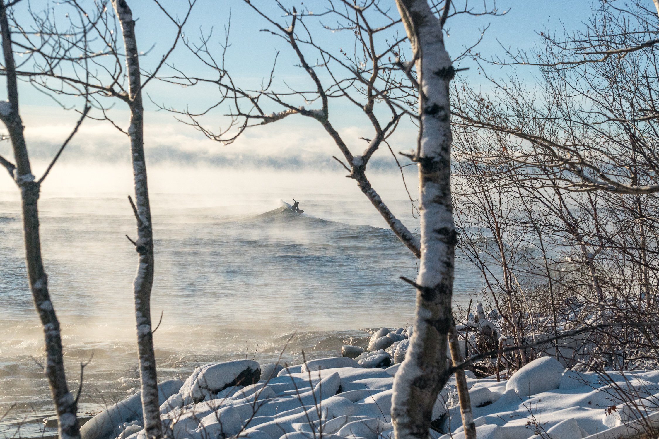 Snow covered shoreline faces out toward fog-covered water with someone surfing off in the distance.